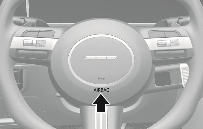 2025 Hyundai Tucson driver's front air bag image. A black arrow points upward to the 'AIRBAG' text located at the bottom center of the steering wheel hub. Indicates the location of the driver's front air bag module.