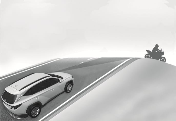 2025 Hyundai Tucson driving on an inclined road image. A white SUV ascends a hill while a motorcycle is positioned over the crest. The camera's detection range, depicted as a shaded cone on the road, fails to detect the motorcycle due to the slope. Explains the limitation of the Forward Collision-Avoidance Assist system on uphill or downhill roads.