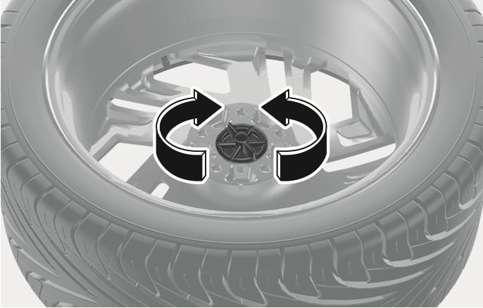 2025 Hyundai Tucson Type B spare tire hold-down bolt image. Large black curved arrows with white outlines surround the central wing bolt, indicating rotation. Explains the operation of turning the bolt to release or secure the spare tire.
