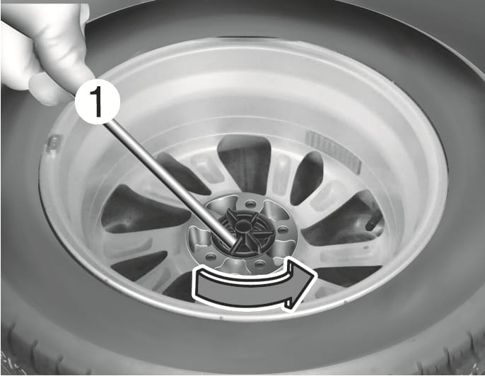 2025 Hyundai Tucson spare tire replacement image. A hand inserts the jack handle (1) into the tire hold-down wing bolt, with a curved arrow indicating counterclockwise rotation. Explains the function of loosening the bolt using the jack handle.