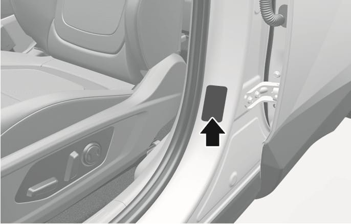 2025 Hyundai Tucson tire specification and pressure label location image. A black arrow points to the rectangular label attached to the driver's side center pillar. The driver's seat and controls are visible for context. Explains the location of the label containing recommended tire pressure information.