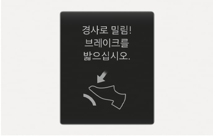 2025 현대 투싼 클러스터 표시창의 경사로 밀림 경고문 이미지입니다. 검은색 배경 화면 중앙에 '경사로 밀림! 브레이크를 밟으십시오.'라는 문구가 표시되어 있습니다. 문구 하단에는 발로 브레이크 페달을 밟는 형상의 아이콘과 페달을 누르는 방향을 지시하는 화살표가 있습니다. 경사로에서 가속 페달로 정차 상태를 유지할 때 변속기 과열을 방지하기 위해 브레이크 페달을 밟도록 안내하는 기능을 설명합니다.