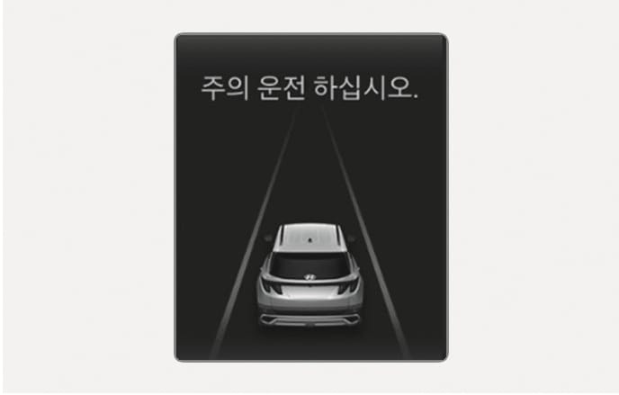 2025 현대 투싼 클러스터 경고문 표시 화면 이미지입니다. 검은색 배경의 화면 중앙에 투싼 차량의 후면 모습과 차선이 그래픽으로 표현되어 있으며, 상단에는 '주의 운전 하십시오.'라는 흰색 문구가 표시되어 있습니다. 긴급 제동으로 정차한 후 제동 제어가 종료될 때 운전자에게 직접 운전할 것을 안내하는 기능을 설명합니다.