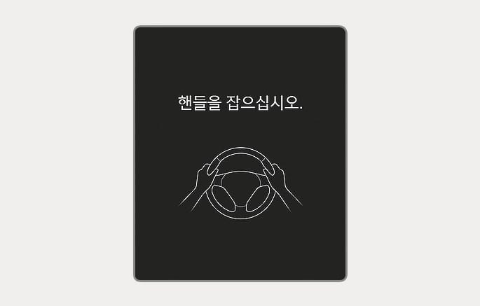 2025 현대 투싼 핸즈오프 경고 이미지입니다. 검은색 배경의 클러스터 화면 중앙에 스티어링 휠을 양손으로 잡고 있는 그래픽이 있고, 그 위에 '핸들을 잡으십시오.'라는 문구가 표시되어 있습니다. 일정 시간 동안 스티어링 휠을 잡지 않을 경우 운전자에게 경고하는 기능을 설명합니다.