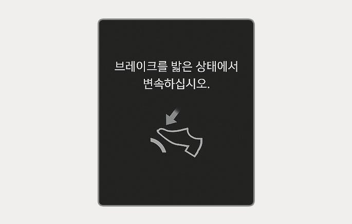 2025 현대 투싼 클러스터 표시창 경고문 이미지입니다. 검은색 배경에 '브레이크를 밟은 상태에서 변속하십시오.'라는 문구가 표시되어 있습니다. 문구 하단에는 발로 페달을 밟는 형상의 아이콘과 아래 방향 화살표가 그려져 있습니다. 브레이크 페달을 밟지 않은 상태에서 변속을 시도할 때 경고 메시지를 띄워 올바른 조작을 안내하는 기능을 설명합니다.